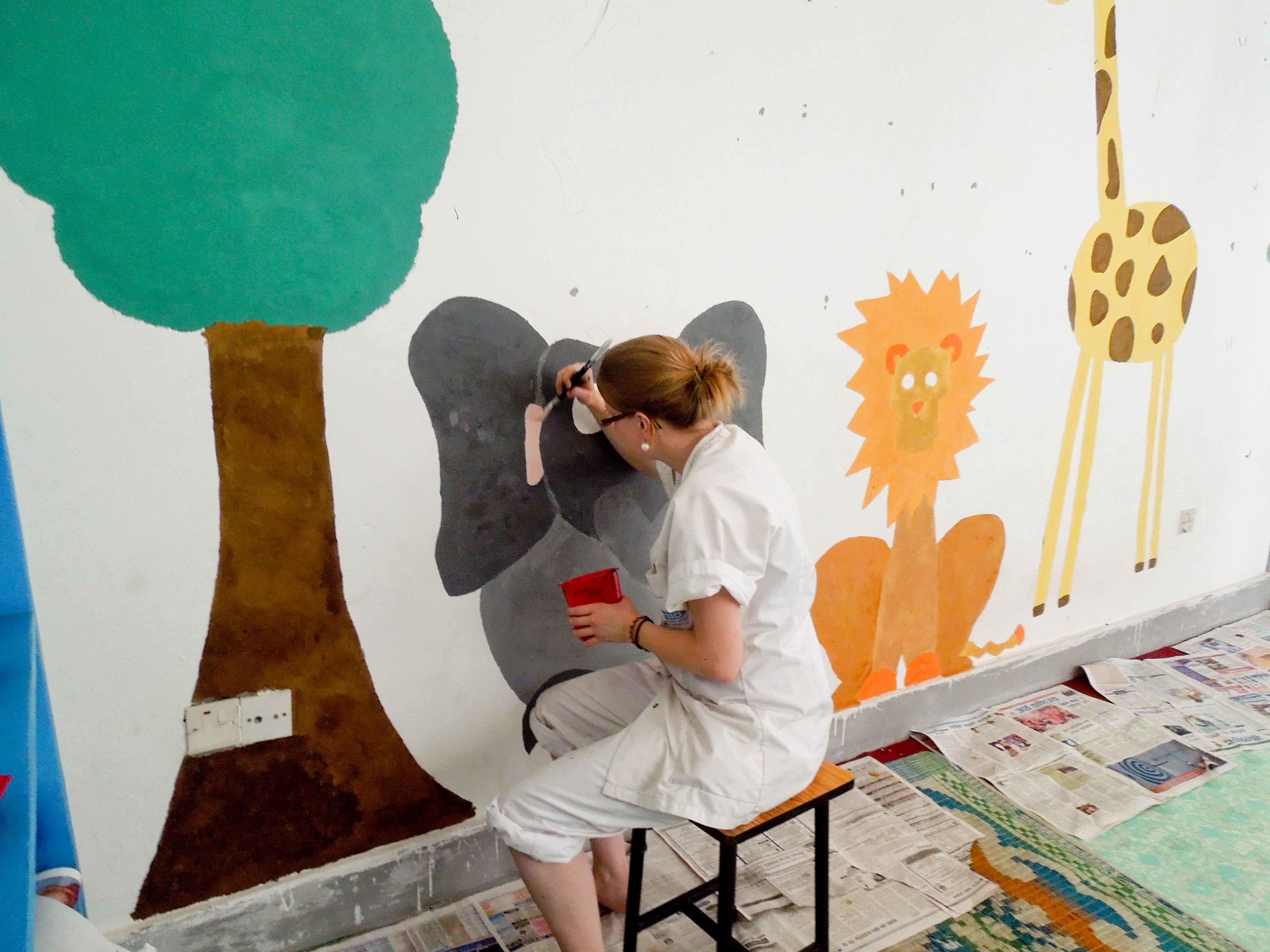 An older adult completing childcare volunteer work in Nepal, paints a mural as part of her project work. A volunteer over 50 doing childcare volunteering in Nepal, paints an educational mural on a wall.